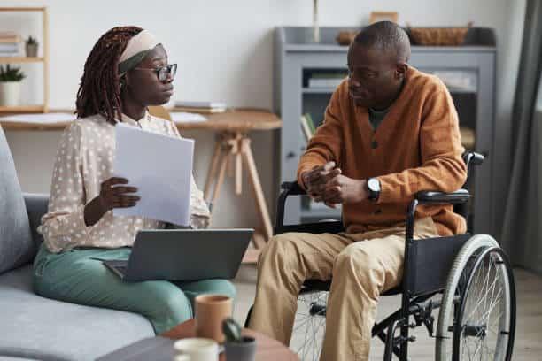 A woman holding documents sits on a couch facing a man in a wheelchair. A laptop is open in front of her. Both are in a living room with a wooden cabinet and table.