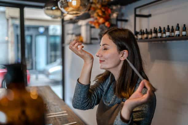 Woman in a gray sweater smelling a perfume strip with bottles on the table and shelves behind her.