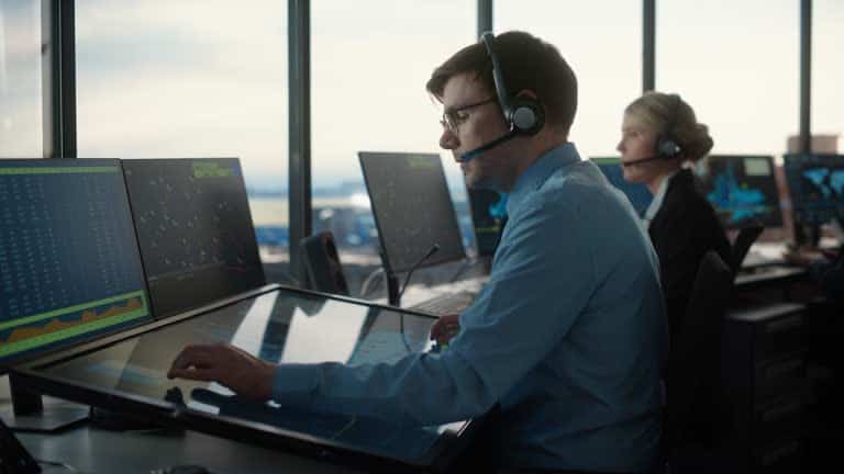 Air traffic controllers focused on large monitors, wearing headsets, in a control room with large windows.