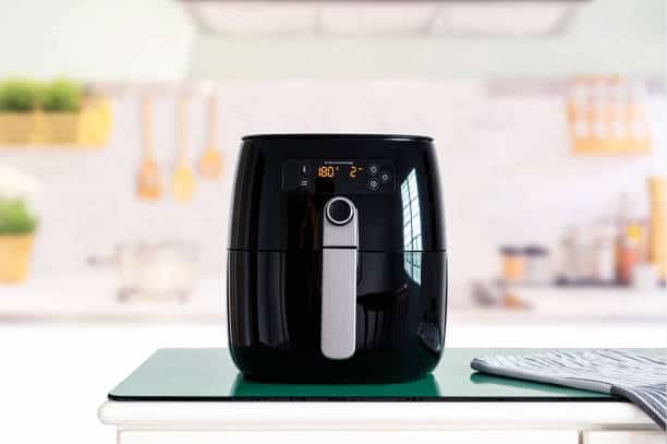 A black air fryer sits on a kitchen counter with a digital display showing 180°C and an image of a fan. A striped pot holder is beside it.