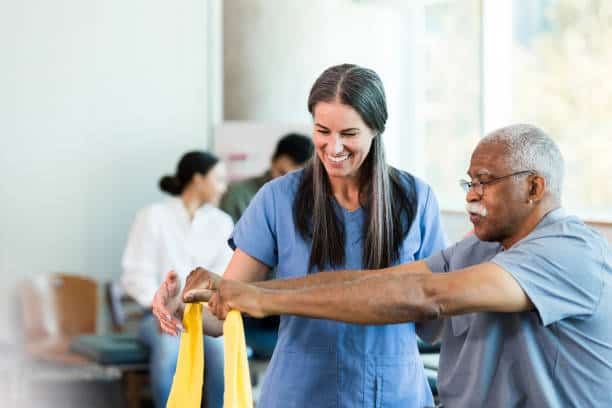 A healthcare professional assists an older man with arm exercises using a resistance band in a clinic setting.