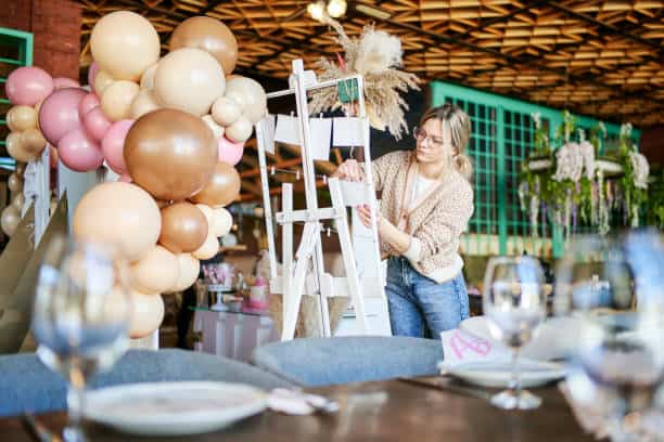 A person adjusts decorations on a stepladder surrounded by balloons in a restaurant. Tables are set with glassware in the foreground.