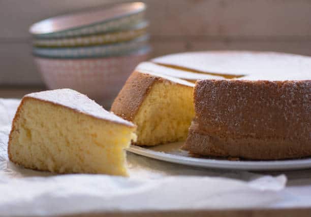 A sliced chiffon cake on a plate with a wedge placed on parchment paper. Stacked bowls are in the background.