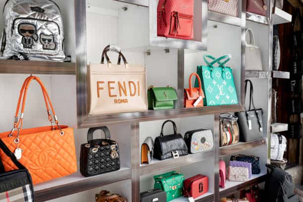 A display of various designer handbags, including Fendi, Louis Vuitton, and Chanel, arranged on white shelving.