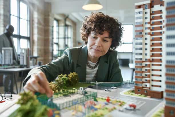 Person examining a detailed architectural model of a residential area with buildings, roads, and trees in an office setting.