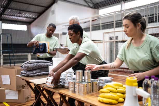 Volunteers organize food and supplies on tables inside a gymnasium, preparing for a distribution event.