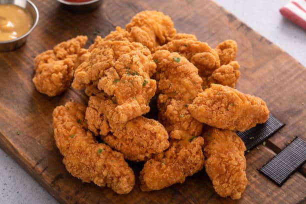 A pile of golden-brown fried chicken tenders on a wooden board with two dipping sauces in small metal bowls nearby.