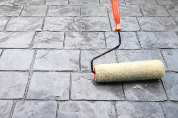 A paint roller with a beige foam covering is being used to seal a gray textured stone tile floor.