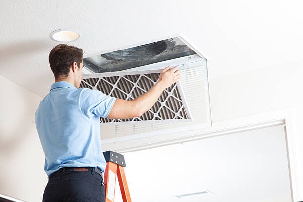 A person on a ladder replaces an air filter in a ceiling vent.