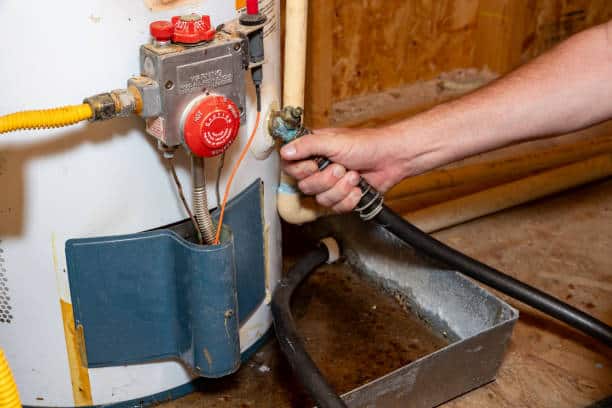 Hand turning a valve on a water heater in a utility room, with visible pipes and a drip pan on the floor.