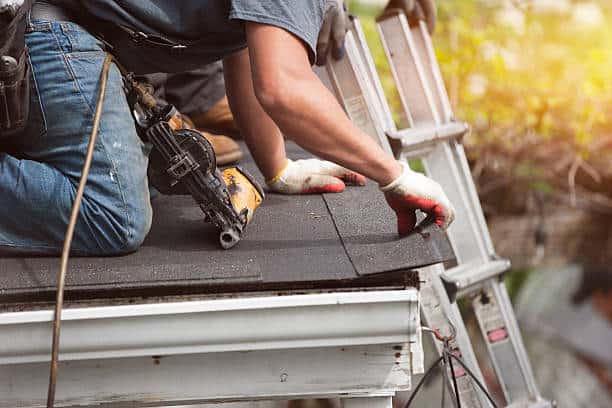 A worker wearing gloves is installing roof shingles while kneeling, with a tool belt and nail gun, near a metal ladder.