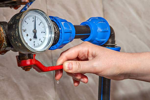 A hand turns a red valve on a blue and black pipe with a pressure gauge, releasing a droplet of water.