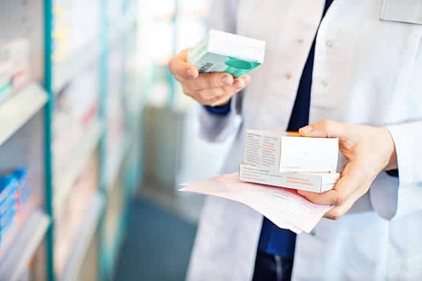 A person wearing a white coat holds prescription boxes and papers in a pharmacy.