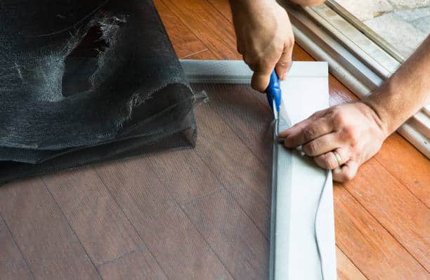 A person repairs a torn window screen using a blue tool on a wooden floor.