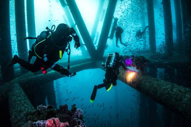 Divers explore an underwater structure surrounded by schools of fish, with beams of light illuminating the scene.