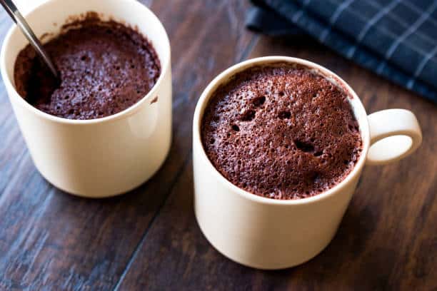 Two white ceramic mugs with chocolate mug cakes on a wooden table, one with a spoon inside.