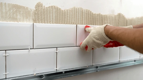 Hands wearing gloves are placing white rectangular tiles on a wall using adhesive and spacers.