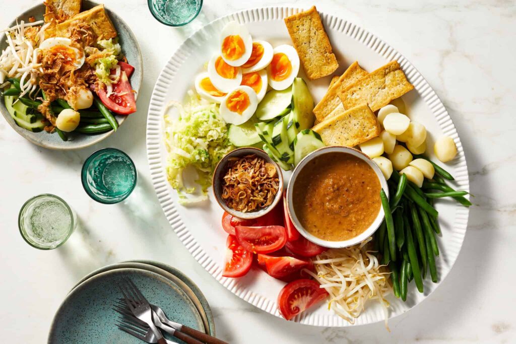 A platter of gado-gado featuring hard-boiled eggs, tofu, vegetables, and peanut sauce, served with a side salad and drinks on a white marble surface.