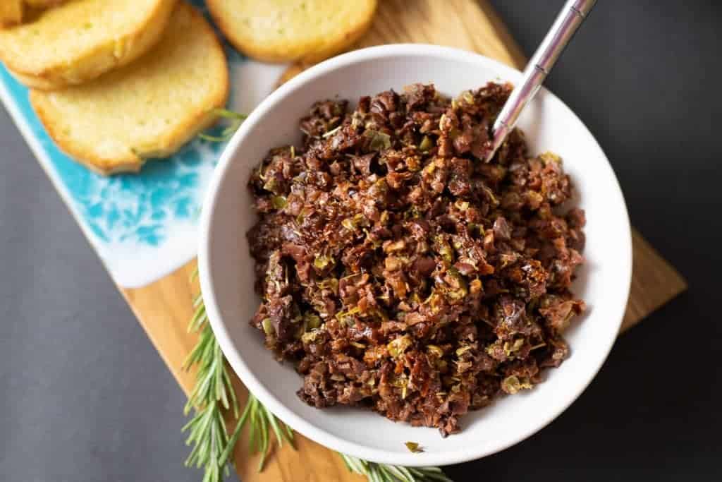 A bowl of olive tapenade with a spoon, on a wooden cutting board alongside slices of toasted bread and a sprig of rosemary.