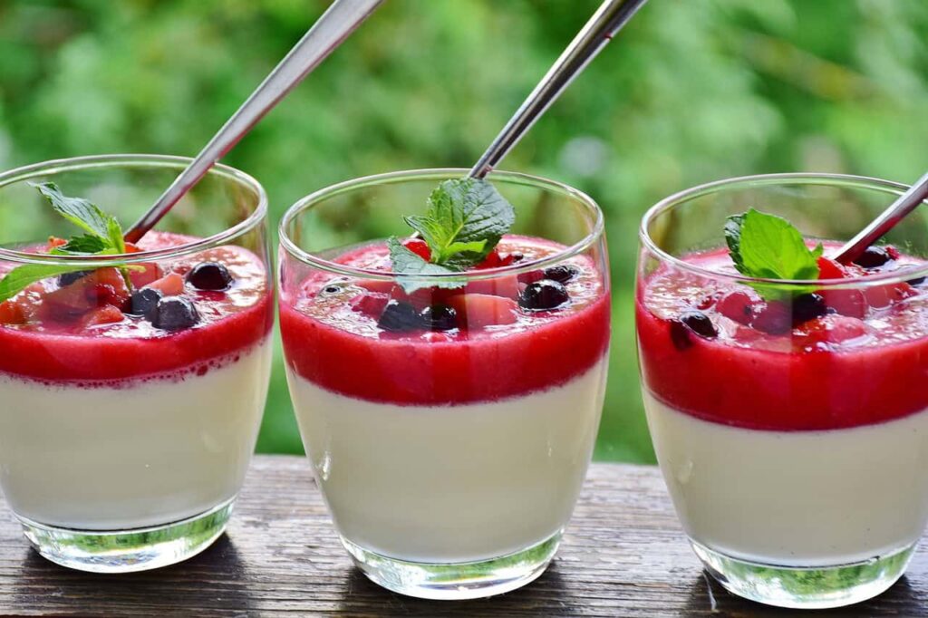 Three glasses of creamy dessert topped with red fruit sauce, blackberries, and mint leaves, each with a spoon, on a wooden surface.