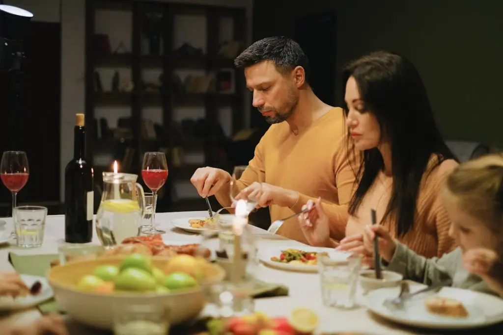 A group of people sitting at a table enjoying a meal, with plates of food, glasses of wine, and a fruit bowl in the foreground.