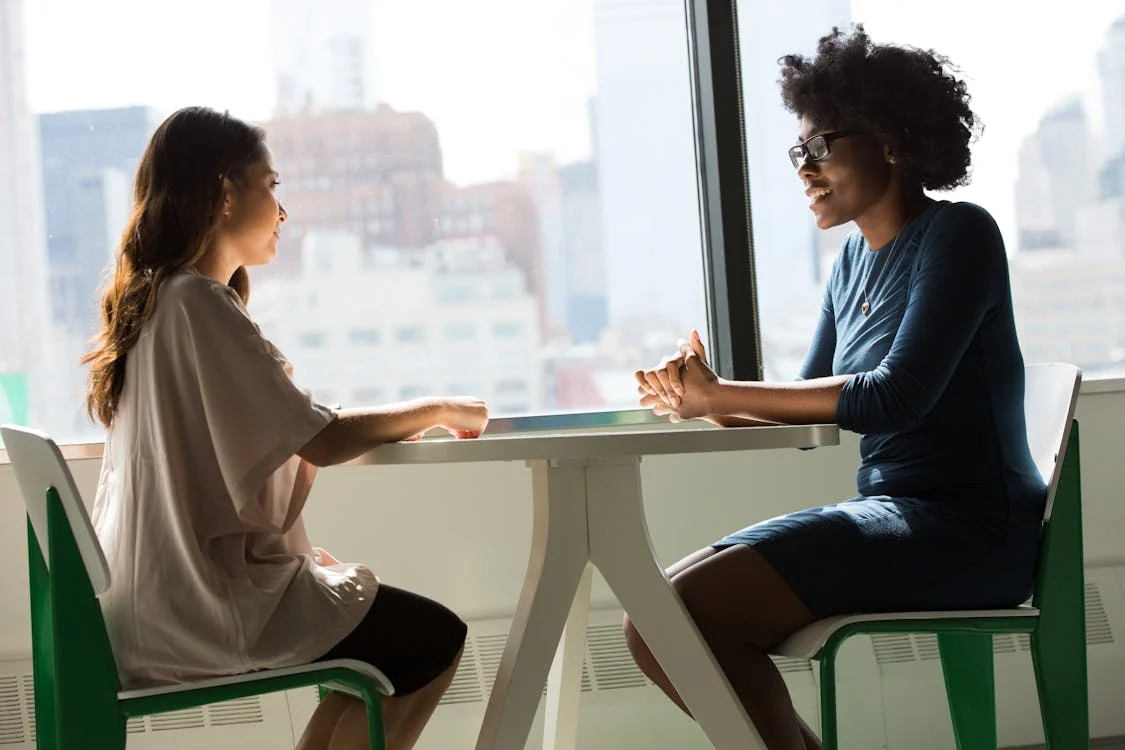 Two people sit across from each other at a table by a window, engaged in conversation, with city buildings visible in the background.