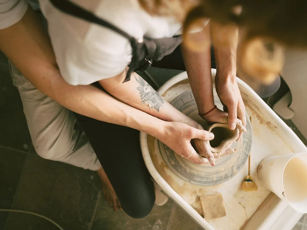 Two people shaping clay on a pottery wheel, with one person's hands guiding the other.