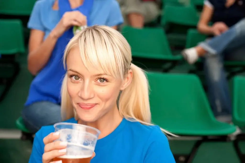A person with blonde hair smiles while holding a plastic cup of beer, sitting in green stadium seats.