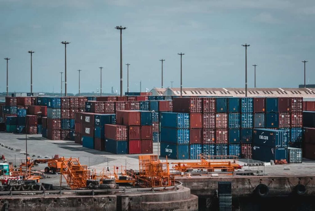 Stacks of shipping containers in a busy port area with heavy equipment and tall light poles.