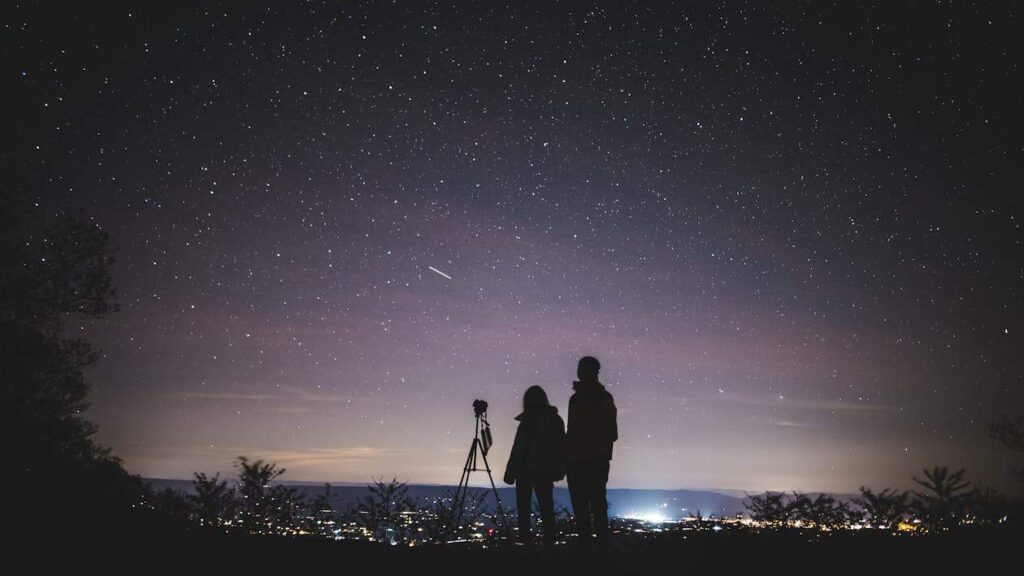 Two people stand by a camera and tripod under a starry sky, overlooking city lights in the distance.