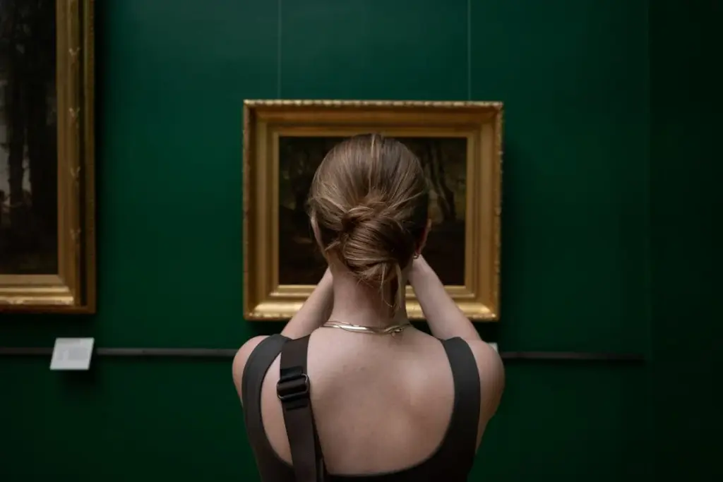 Person with a neat bun hairstyle stands in front of a framed painting on a green wall, viewed from behind.