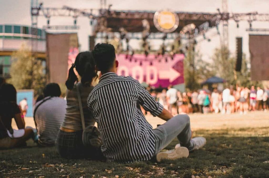 People sit on the grass at an outdoor event with a stage in the background, featuring a large screen and a crowd.