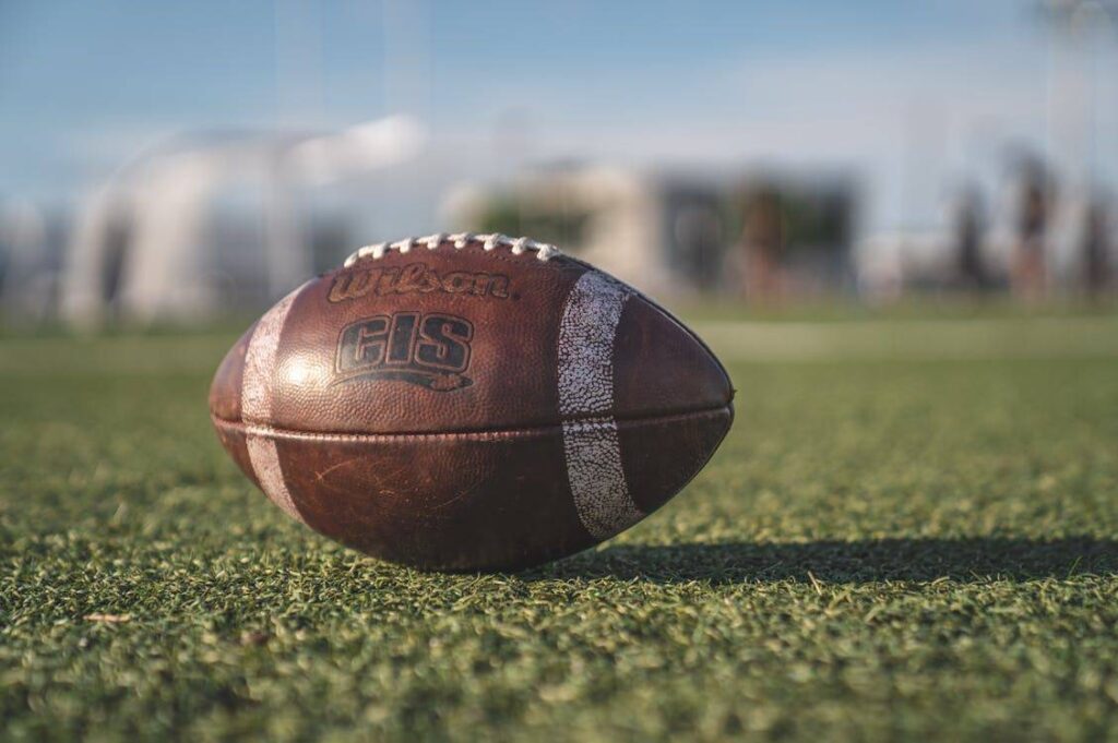 Close-up of a Wilson GIS American football on a grassy field, with blurred structures and sky in the background.