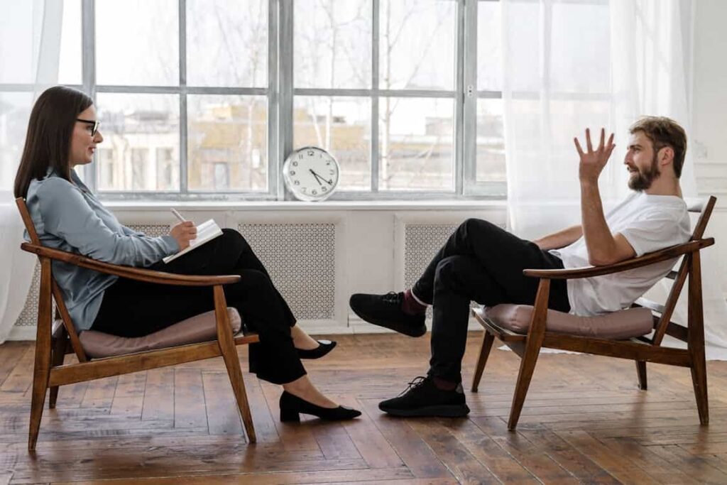 A woman and a man sit facing each other in chairs, engaged in conversation in a room with wooden floors and a window. A clock is on a table between them.