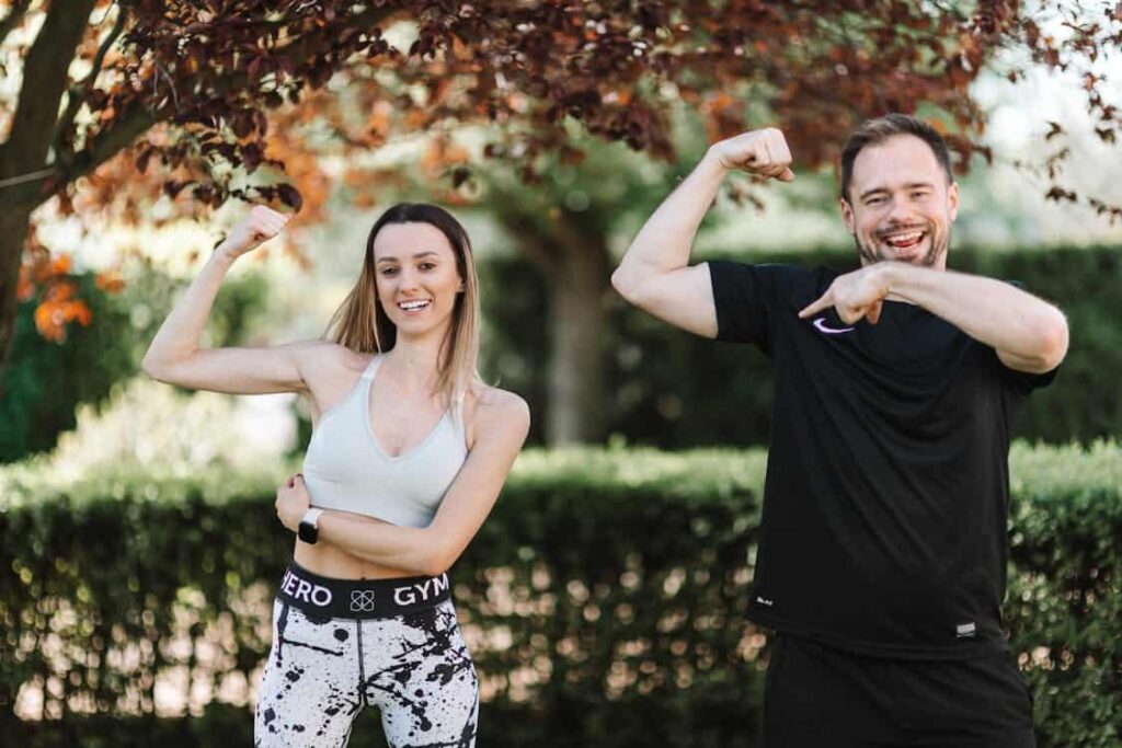 A man and a woman flexing their muscles and smiling outdoors, standing in front of trees and greenery.