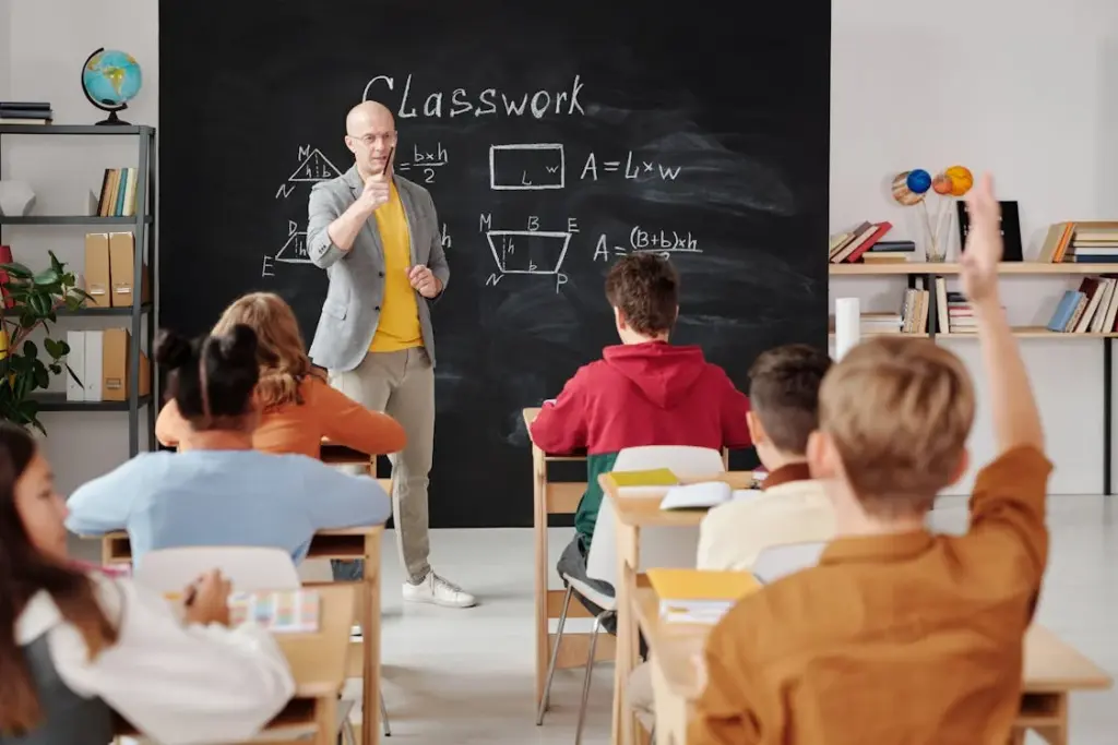Teacher explaining geometry to students in a classroom with a blackboard displaying formulas and diagrams.