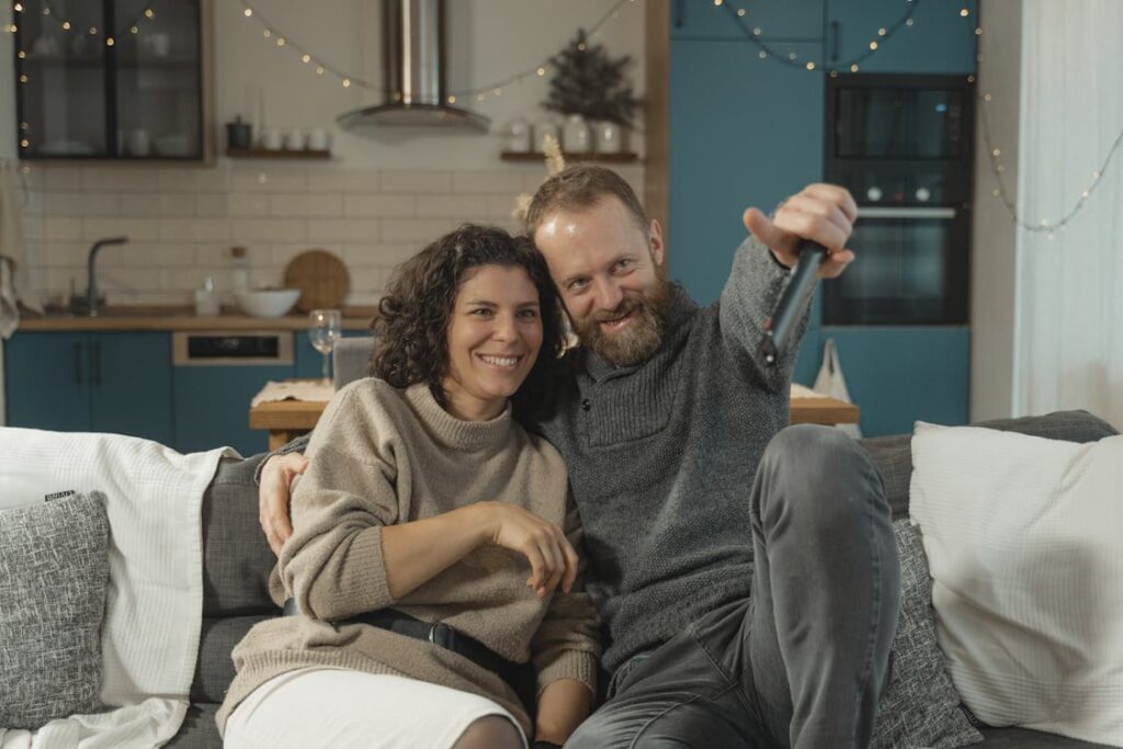 A couple sits on a sofa in a cozy living room, smiling at the camera. The man is holding a TV remote.