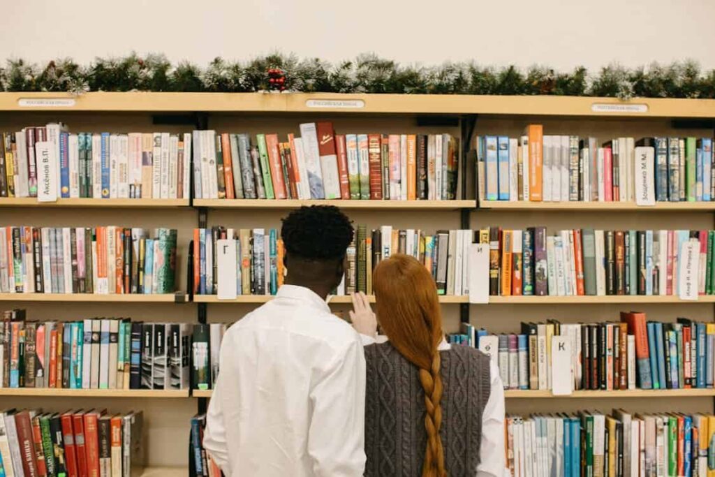 Two people stand in front of a bookshelf, with one pointing at books. A festive garland decorates the top shelf.