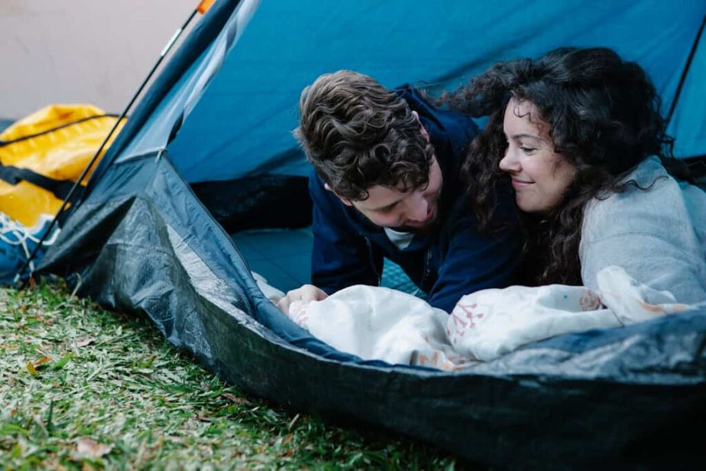 Two people lying inside a tent, wrapped in blankets, smiling at each other.