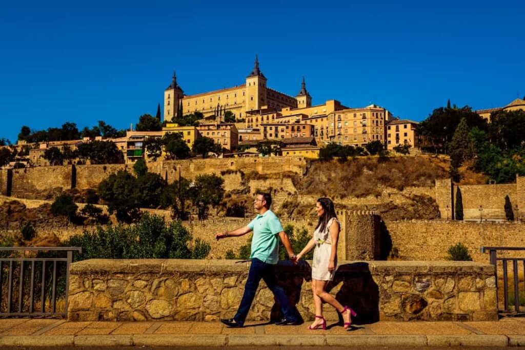 A man and woman holding hands walk along a stone path with a historic building and cityscape in the background under a clear blue sky.
