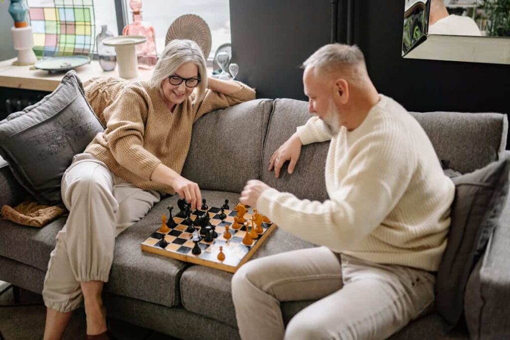 Two people sitting on a couch playing chess, with a chessboard between them. Cozy room with cushions and decorative items in the background.