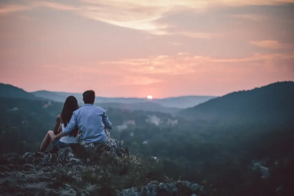 A couple sits on a rocky hill overlooking a landscape at sunset, with mountains and a colorful sky in the background.