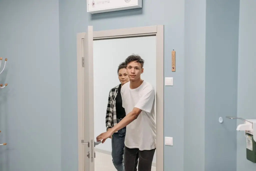 Two people are entering a room through a door, with the person in front holding the door handle. The room has blue walls and a coat rack on the left.