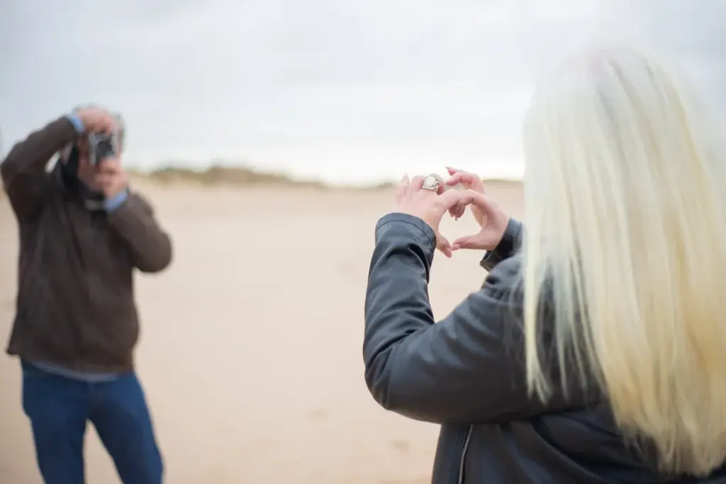 A person with blonde hair forms a heart shape with their hands on a beach, while another person in a brown jacket takes a photo.