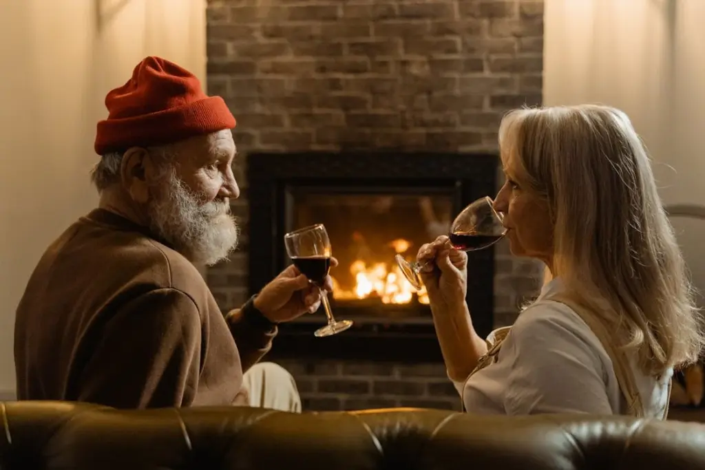 An older man and woman sit on a couch, enjoying glasses of red wine in front of a lit fireplace.
