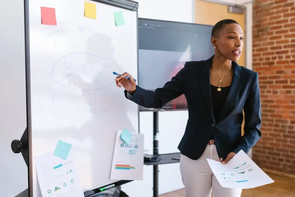 Person presenting a budget plan on a whiteboard, pointing with a pen, holding printed charts.