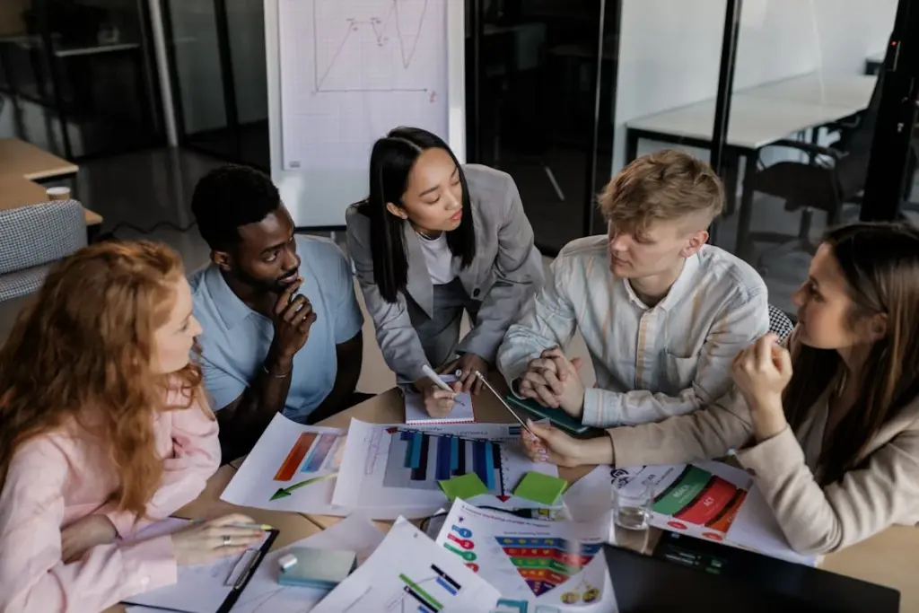 Five people in a meeting room are gathered around a table with charts and documents, engaged in discussion.