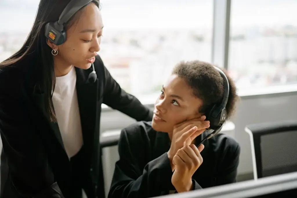 Two professionals in a call center wearing headsets, engaged in a conversation, with large windows in the background.