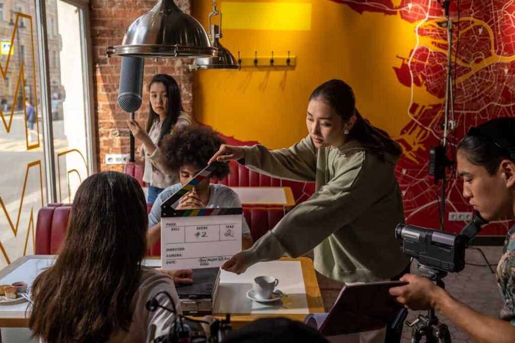 A film crew records a scene in a café, with a director, a clapperboard, and a boom microphone visible, as actors sit at a table with a coffee cup.
