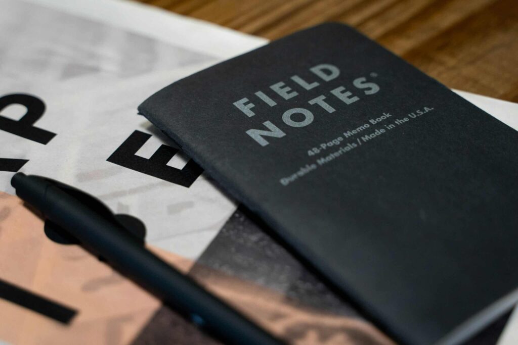 A black Field Notes notebook and pen on top of scattered papers.